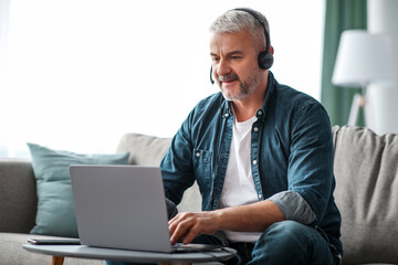 Successful handsome bearded mature man having video business call from home, using headset and laptop, sitting on couch in living room, talking to business partners, smiling and gesturing, copy space