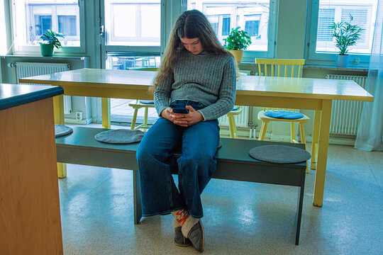 Young woman sitting with poor posture looking down at her smartphone in casual kitchen setting. Extended phone use.  Unhealthy digital habits of modern living.

