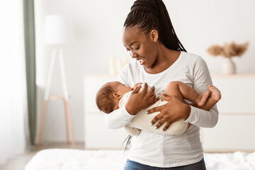 Maternity Concept. Portrait of smiling African American woman holding her adorable small black upset baby on hands, singing lullaby to sleep, comforting infant kid standing in bedroom at home