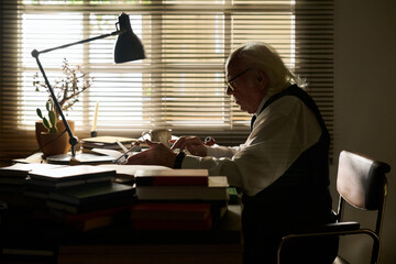 Senior Caucasian man sitting at desk writing on paper, surrounded by stacked books and office supplies, concentrating on work in front of window with blinds partially closed