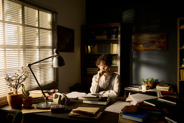 Middle aged Caucasian woman sitting at desk reading documents in home office surrounded by books...