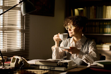 Caucasian middle aged woman sitting at desk holding cup and saucer, wearing glasses, looking out...