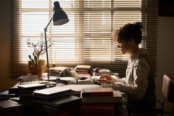 Caucasian young adult woman sitting at desk typing on vintage typewriter surrounded by stacks of...