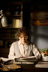 Portrait of young adult Caucasian woman wearing glasses sitting at desk using typewriter, focusing...