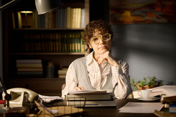 Portrait of middle aged woman sitting at desk resting chin on hand looking into camera surrounded...