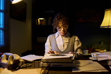 Caucasian young adult woman sitting at desk working on typewriter surrounded by scattered papers...