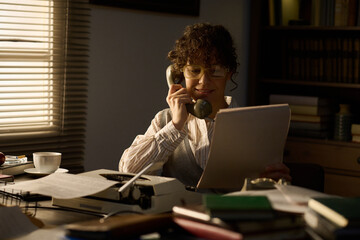 Caucasian middle aged woman sitting at desk holding telephone receiver while reading document,...