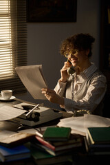 Caucasian young adult woman sitting at desk talking on landline phone while holding document,...