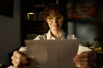 Portrait of Caucasian middle aged woman smiling while holding paper with both hands, sitting at...