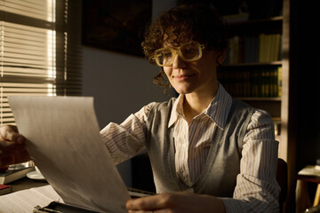 Caucasian middle aged woman sitting at desk reading document with slight smile, wearing glasses and...