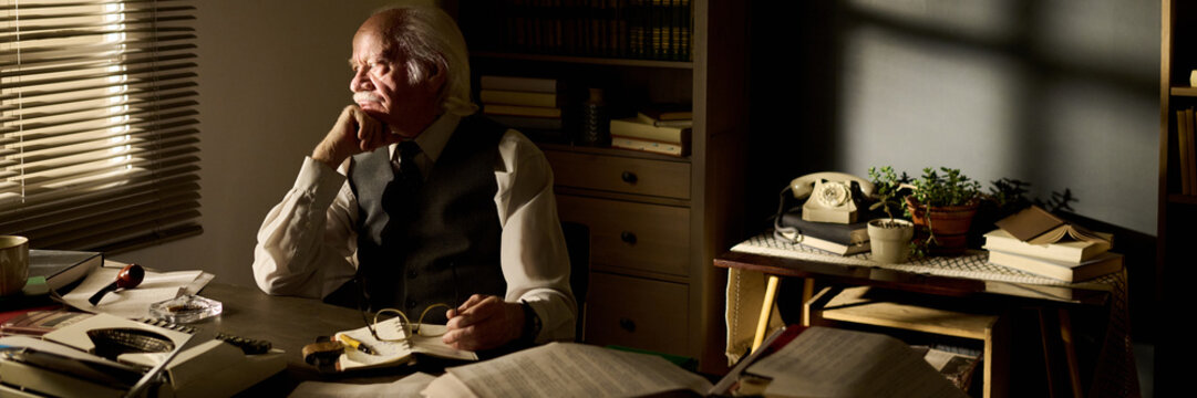 Senior Caucasian man sitting at desk holding eyeglasses, gazing out window with thoughtful expression, surrounded by open books and vintage telephone in office setting - Powered by Adobe