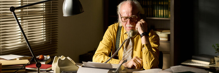Senior Caucasian man sitting at desk talking on landline telephone, wearing glasses and...