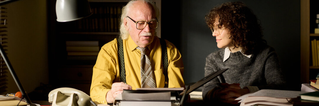 Senior Caucasian man wearing glasses discussing documents with young adult Caucasian woman at desk, both sitting and engaging in conversation surrounded by paperwork and books
