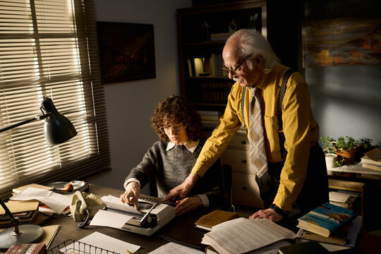 Caucasian middle aged woman using typewriter while senior Caucasian man standing beside her pointing at document, both focused on paperwork in office setting with books and files