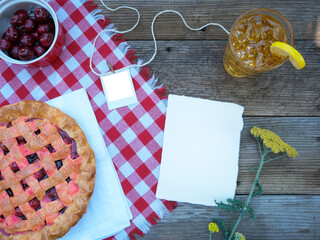 Overhead blank invitation note paper, transparent photo frame, and cherry pie on red checkered...