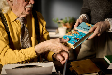 Senior Caucasian man sitting at desk receiving book from young adult Caucasian woman, both focusing on exchanging colorful travel-themed book titled My Journey
