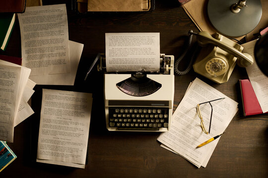 Vintage typewriter sitting on wooden desk surrounded by scattered typed manuscript pages, rotary telephone, eyeglasses, pen, and books, evoking classic writing workspace