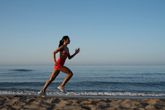 Energetic fit woman in red sportswear running along the beach at sunrise, embracing healthy lifestyle and outdoor cardio workout with focus and determination by the sea
