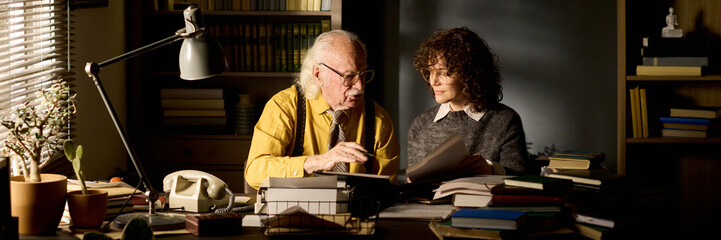 Senior Caucasian man explaining documents to young adult Caucasian woman at desk, both sitting...