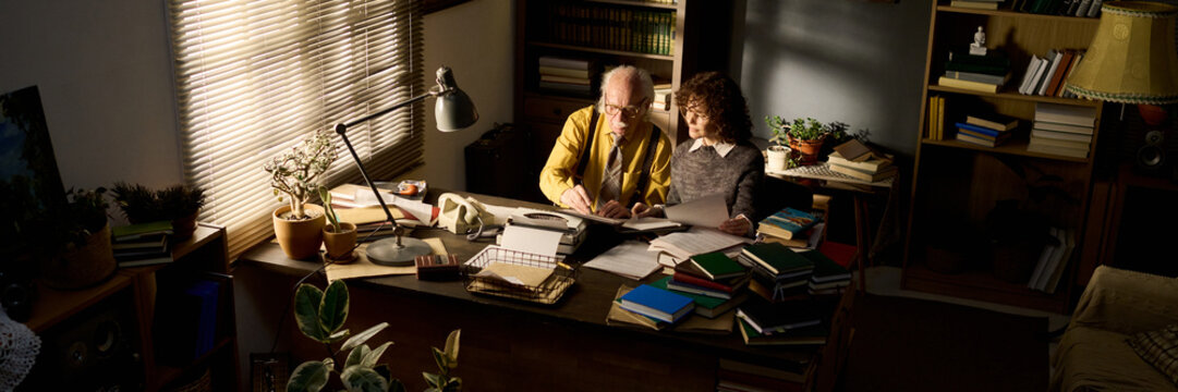 Senior Caucasian man sitting at desk helping young Caucasian boy with homework, both looking at open books surrounded by stacks of papers and books in home office setting - Powered by Adobe