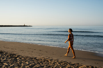 Fit young woman walking barefoot on sandy beach during sunrise, wearing red bikini, looking ahead...
