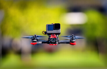 Flying FPV racing drone front-facing with an action camera on top. The quadcopter is hovering in a park with trees