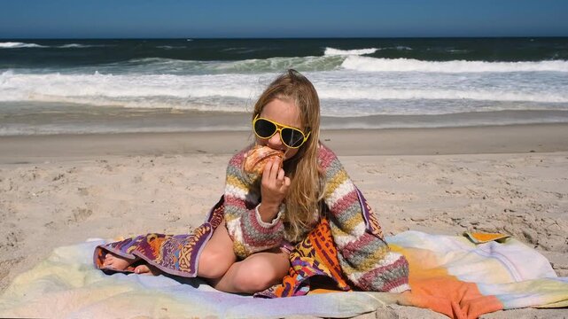 A child wrapped in a colorful towel eats a croissant while sitting on the sand near the Atlantic Ocean. The scene captures a relaxed beach moment with waves in the background