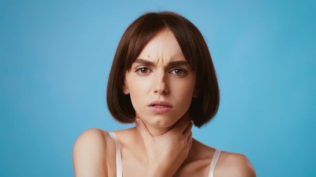 A young brunette woman looks directly at the viewer with a thoughtful expression. She is touching her neck, suffering from tonsilitis
