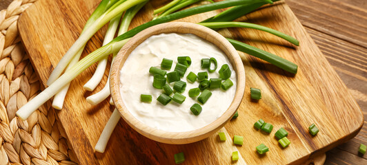 Board and bowl of tasty sour cream with sliced green onion on wooden background