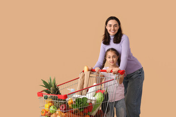 Beautiful young happy mother and her daughter with full shopping cart on beige background