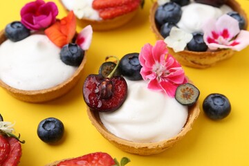 Sweet tartlets with berries and flowers on yellow background, closeup. Delicious dessert