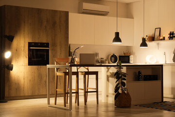 Interior of modern kitchen with counters, table and glowing lamps at night