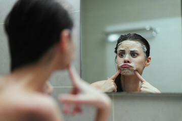Pensive young woman applying face mask in bathroom, looking at herself in the mirror while making funny facial expressions. Self-care and beauty routine concept.