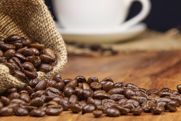A jute bag with roasted coffee beans scattered on a rustic wooden table, next to it is a cup of freshly brewed coffee