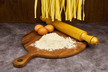 Fresh egg fettuccine noodles, drying in the foreground, hanging over a wooden table containing flour and egg, basic ingredients for preparation.