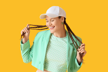 Stylish young woman with dreadlocks and cap on yellow background