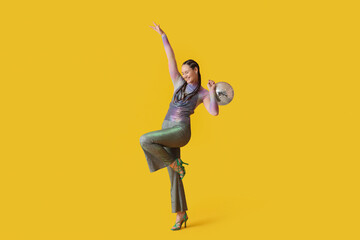 Fashionable young woman with dreadlocks and disco ball dancing on yellow background