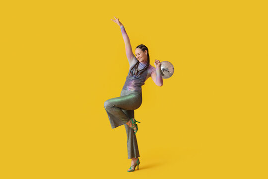 Fashionable young woman with dreadlocks and disco ball dancing on yellow background