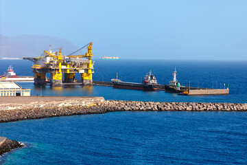 Large offshore oil platform docked at an industrial port, with tugboats assisting in calm deep...