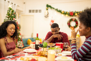 A family enjoys a festive Christmas dinner, sharing food and creating memories. A beautifully decorated Christmas tree and wreath add warmth, while candles and decor enhance the cozy holiday setting.