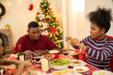 A joyful family shares a Christmas meal together, passing food and creating holiday memories. Decorated tree, festive lights, and presents in the background add warmth to cozy celebration atmosphere.