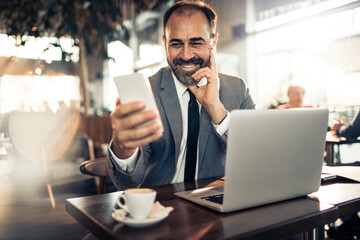 Young businessman holding smartphone at cafe table with laptop
