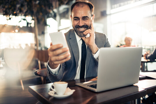 Young businessman holding smartphone at cafe table with laptop - Powered by Adobe