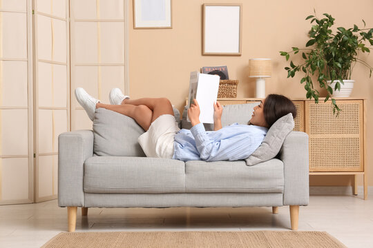 Young Asian woman reading magazine on grey sofa at home