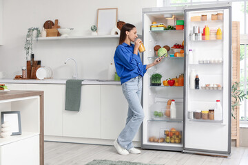 Young woman with juice using mobile phone near open fridge in kitchen
