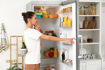 Thoughtful young woman near open fridge in kitchen