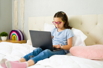 Little girl doing her homework on laptop while relaxing in bed