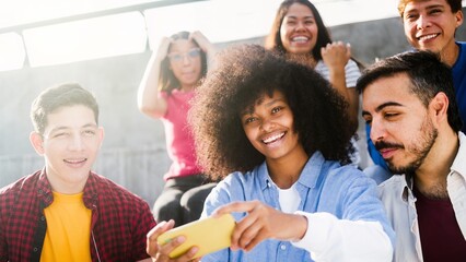 Diverse group of friends sitting in urban park using mobile phone together at sunset, multiracial happy moment