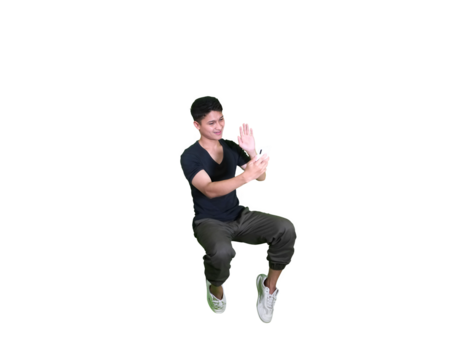 A young man in casual attire is captured mid-air in a squatting pose, looking down at his hands against a white background.