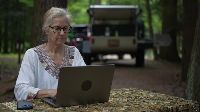 A retired woman happily uses her laptop and smartphone at a campground, with her teardrop camper trailer in the background surrounded by trees.